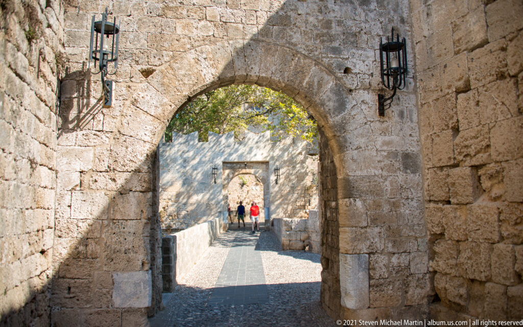Stone archway framing a second gate and two visitors along a sunlit fortified passage.