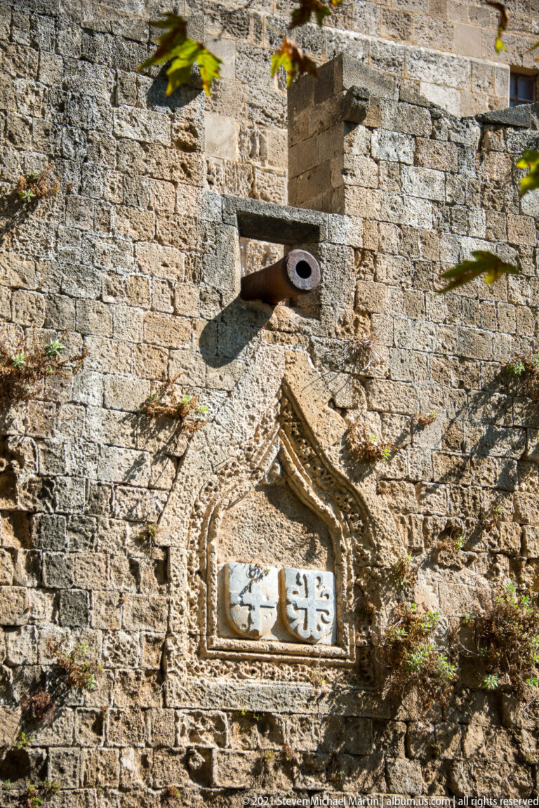 Weathered heraldic shield in a pointed stone frame beneath a projecting metal spout on a fortification wall.