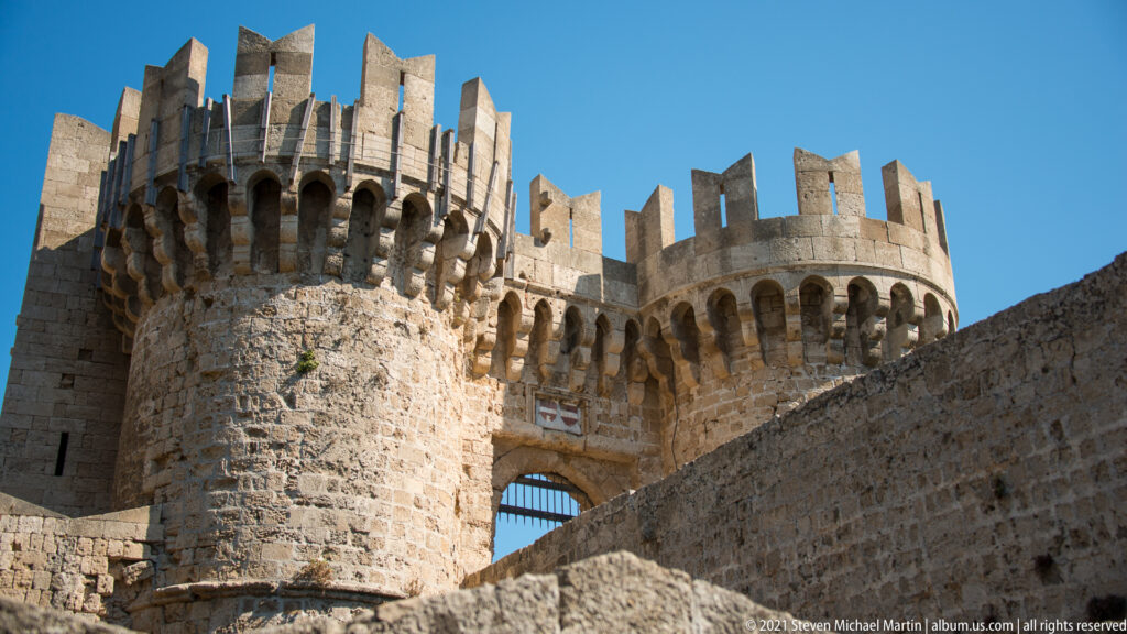 Low-angle view of twin rounded stone towers with corbels, tall crenellations, and a gateway between them.