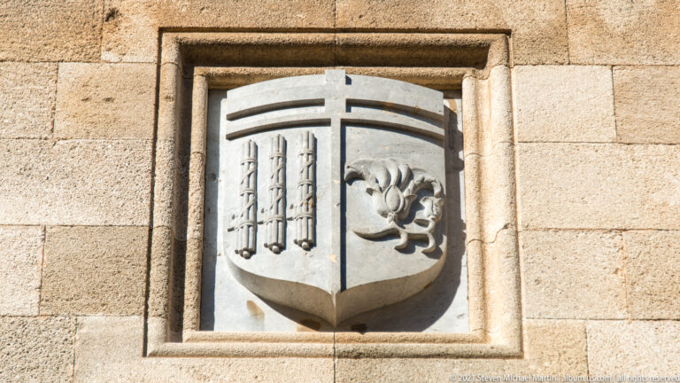 Carved stone shield divided between vertical bars and a floral emblem within a rectangular wall recess.