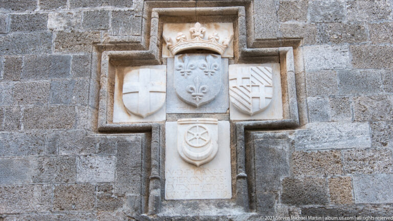 Carved heraldic ensemble with crowned fleur-de-lis shield, surrounding coats of arms, and an inscription below.