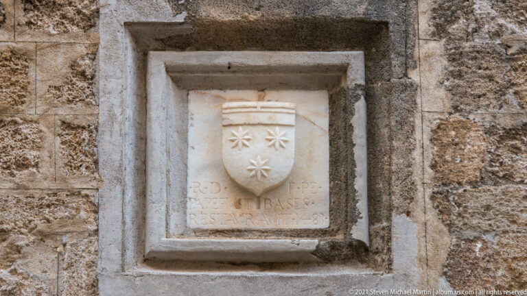 Small stone shield with three star-like motifs above a weathered inscription in a rectangular recess.