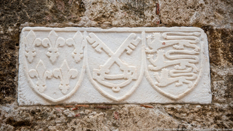 Three carved stone shields showing fleur-de-lis, crossed devices, and striped motifs on a masonry wall.