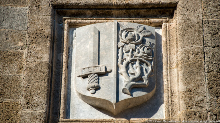 Split stone shield with a sword-like emblem marked FERT and a deeply carved rose motif.