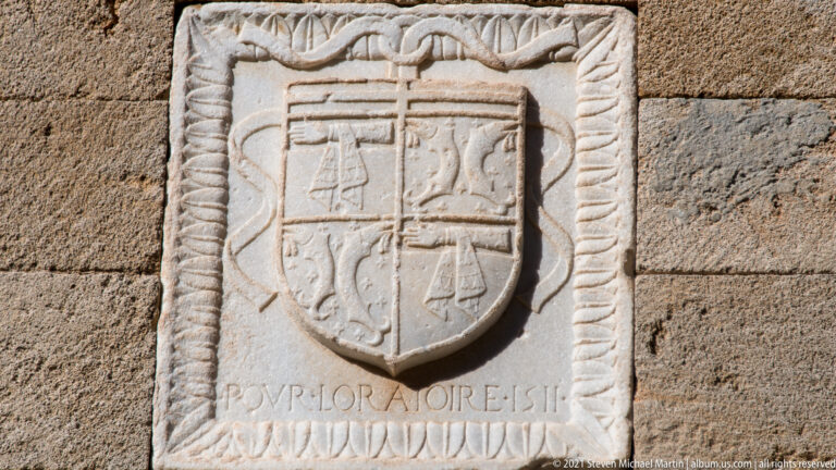 Quartered stone heraldic shield framed by rope-like carving and leaf ornament above a worn inscription.