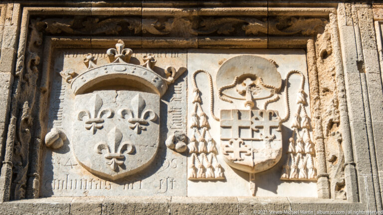 Two ornate heraldic shields, including crowned fleur-de-lis arms and another beneath an ecclesiastical hat with tassels.
