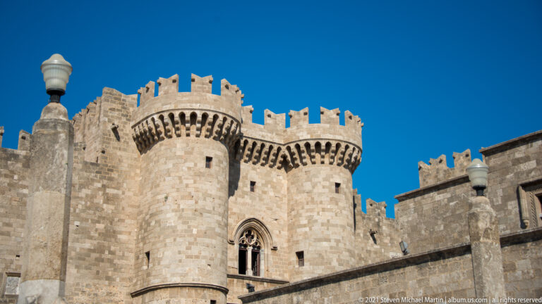 Round crenellated limestone tower with projecting corbels beside a pointed Gothic window under a deep blue sky.