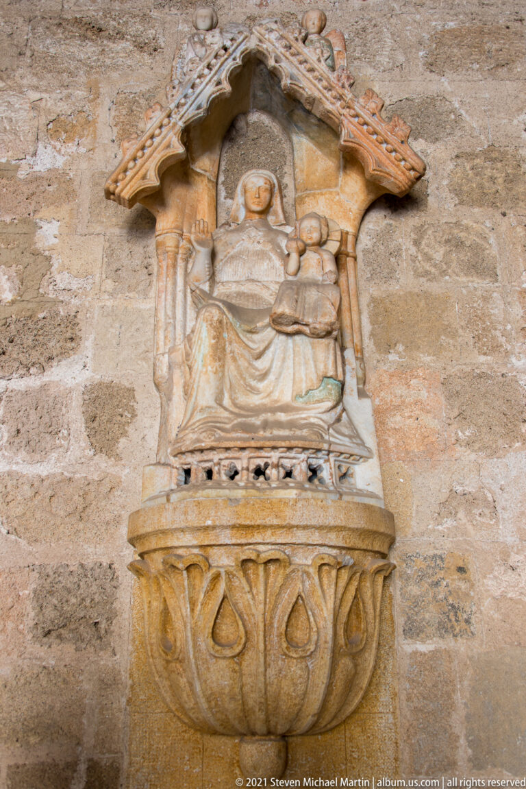 Stone relief of an enthroned woman holding a child beneath a small canopy, mounted above a carved projecting stone element.