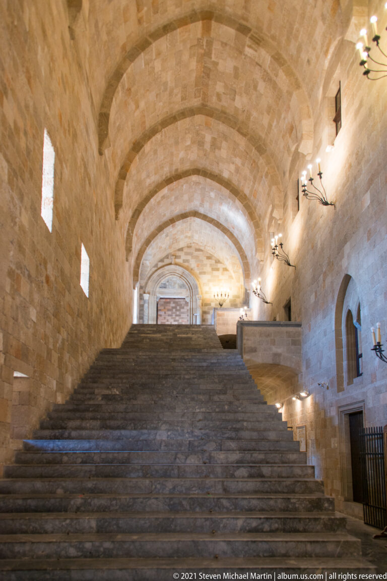 Centered view up a broad stone staircase beneath a long vaulted ceiling, with small windows on the left.