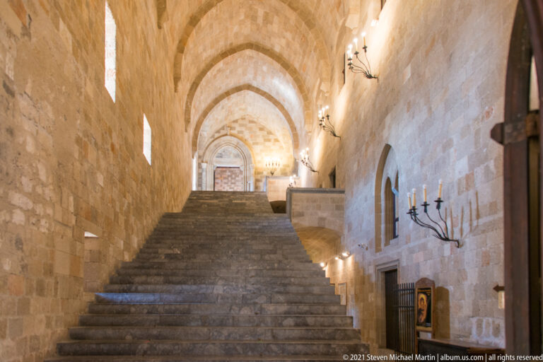 Stone staircase under repeated vaults, viewed beside an arched doorway and black iron wall-mounted candle fixture.