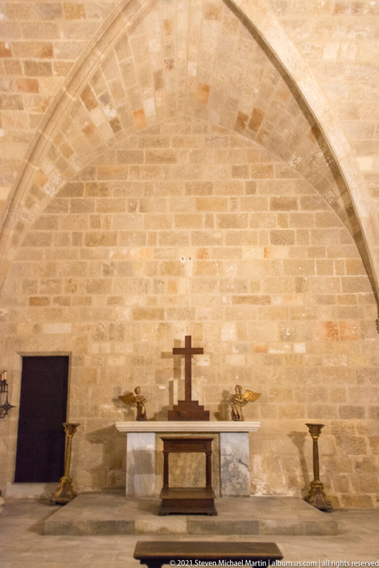 Small altar with a dark cross and candlesticks against a limestone wall beneath a pointed stone vault.