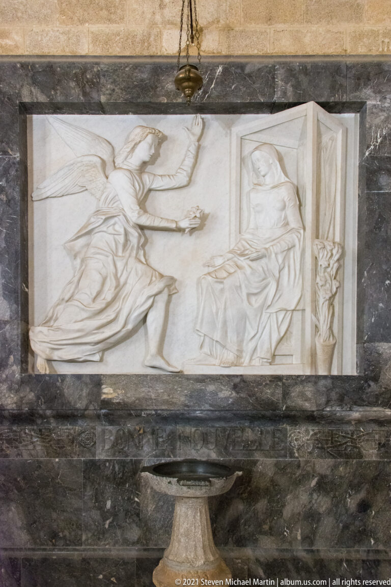 White marble relief showing two draped figures, one standing or approaching and one seated, in a dark stone wall surround.