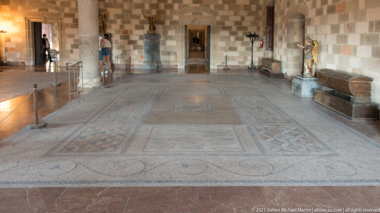 Long stone gallery with a large panelled mosaic floor, sculptures along the walls, and doorways aligned at the far end.