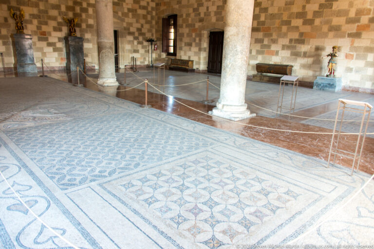 Large geometric mosaic pavement in a stone gallery with a central column and sculptures displayed around the walls.