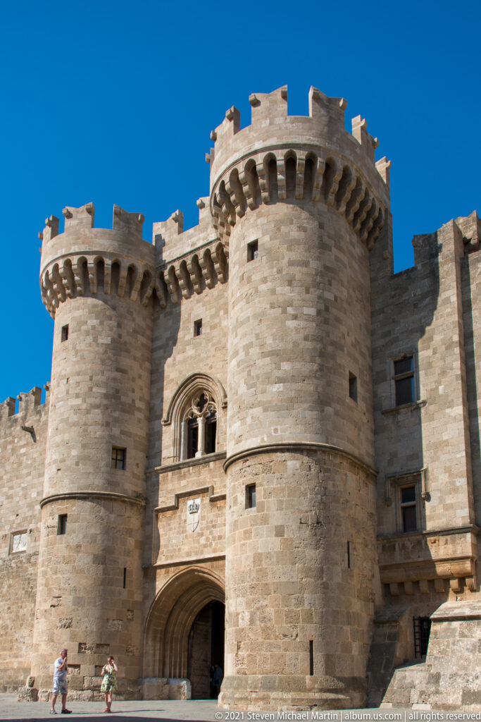 Twin round crenellated towers flank a recessed pointed entrance arch, with a Gothic window centered above the gateway.
