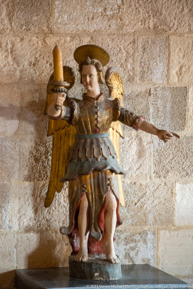 Painted winged angel sculpture with halo, gilded wings, blue-and-gold costume, and a candle holder in one hand.
