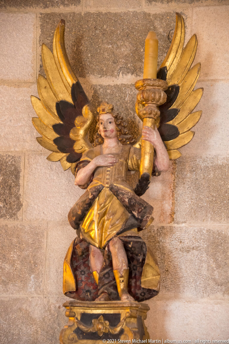 Seated painted angel with broad gilded wings holding a large upright candle socket against a stone wall.