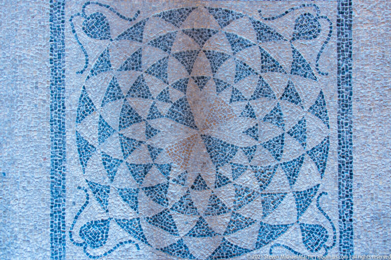 Square floor mosaic with a radiating pinwheel-like geometric pattern in dark, terracotta, and pale tesserae.