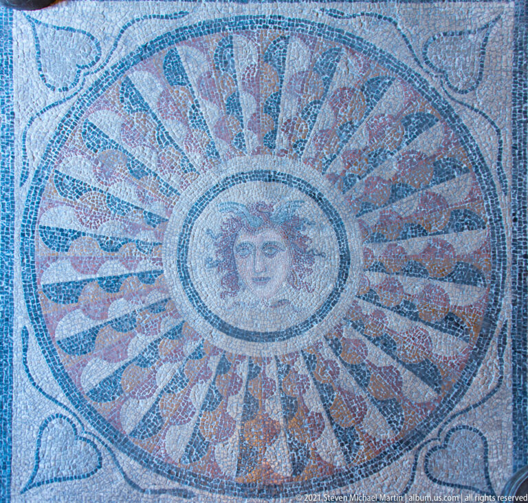 Circular mosaic with a woman’s face at the center of a radiating red, dark, and pale patterned medallion.