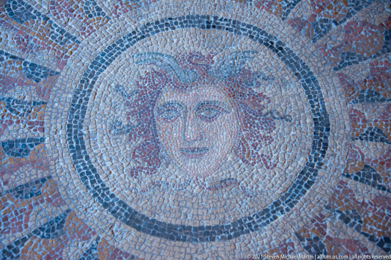 Close-up of a female face in a circular mosaic medallion, with wavy hair and concentric tessellated borders.