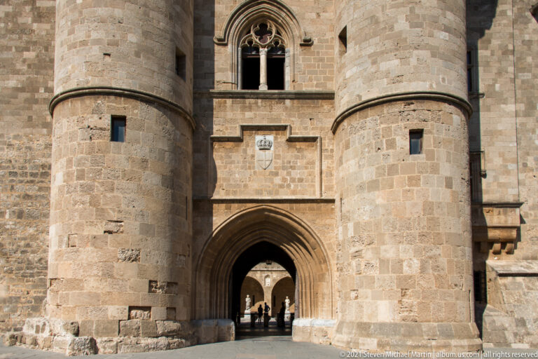 Pointed arched gateway between two massive round stone towers, with several people visible in the passage beyond.