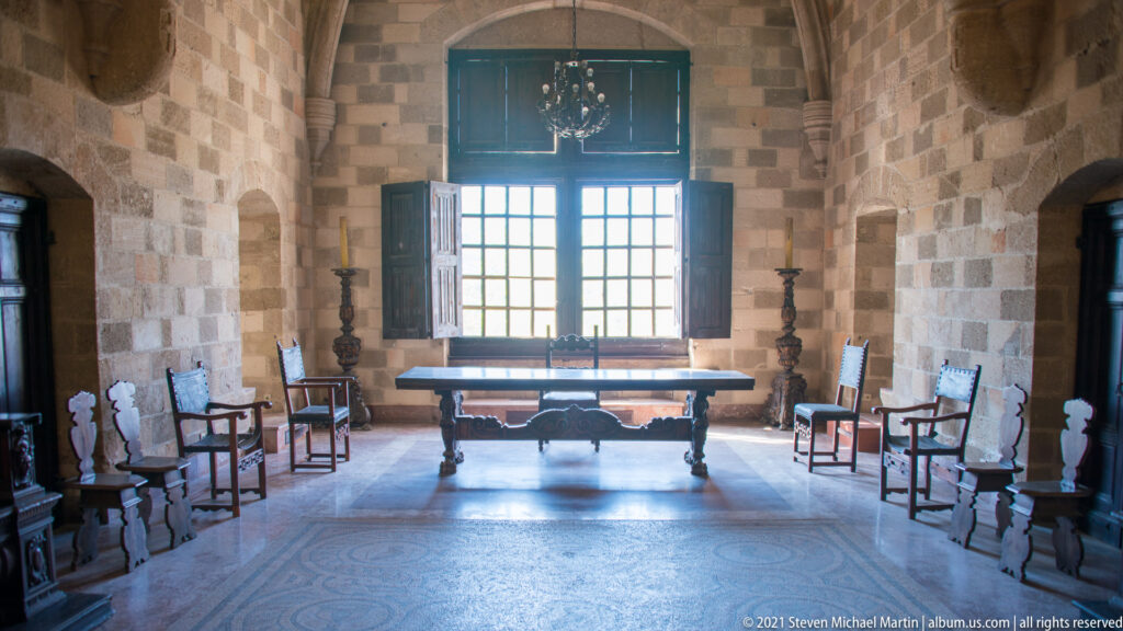Formal stone room with a long central table, high-backed chairs along both walls, and a tall bright shuttered window.