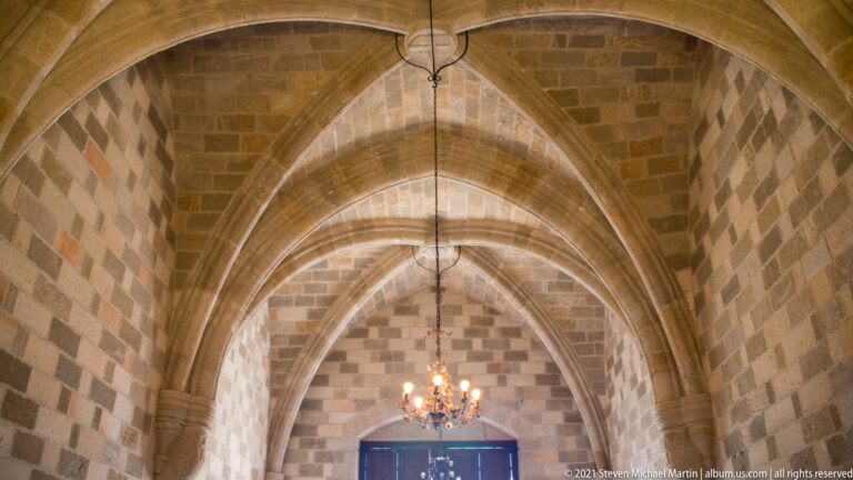 Long stone hall with pointed rib vaults, checker-pattern masonry walls, hanging chandelier, and doorway at the far end.