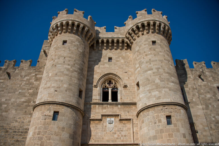 Low-angle view up two cylindrical crenellated towers with a pointed traceried window and shield between them.