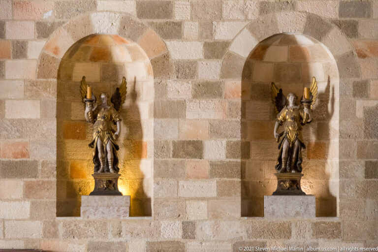 Two winged armored metal figures stand on pedestals in adjacent arched niches set into checker-pattern stone masonry.