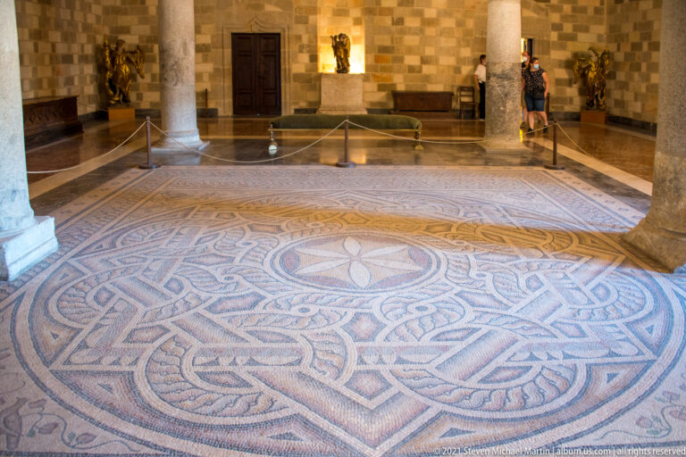 Large geometric mosaic pavement with central rosette and interlace patterns inside a stone hall with columns, barriers, and visitors.