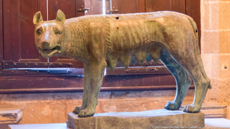 Standing metal she-wolf sculpture with lowered head and visible teats on a stone base before dark wooden doors.