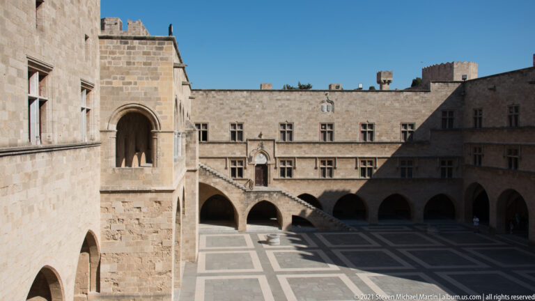 Sunlit stone courtyard with arcades, exterior staircase, rows of windows, and a projecting arched opening on the near wing.
