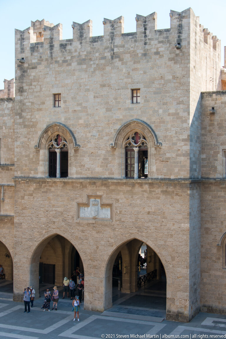Crenellated stone courtyard façade with two arched upper windows, broad ground-level arcades, and visitors seated below.