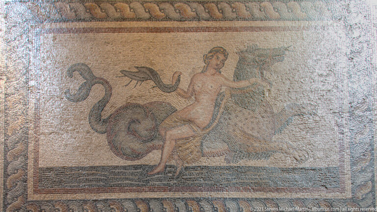 Ancient mosaic showing a nude male figure riding or grappling with a large sea creature within a patterned border.