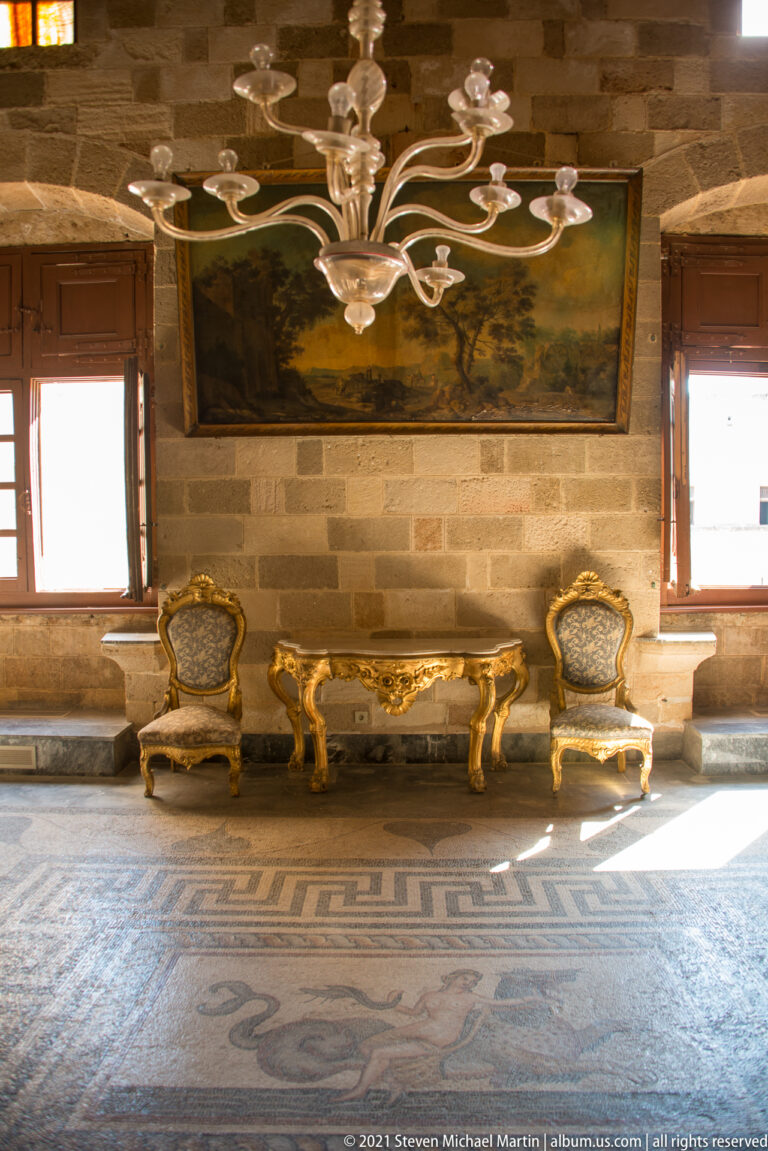 Gilded chairs and table beneath a large landscape painting in a stone room with chandelier, wooden windows, and mosaic floor.