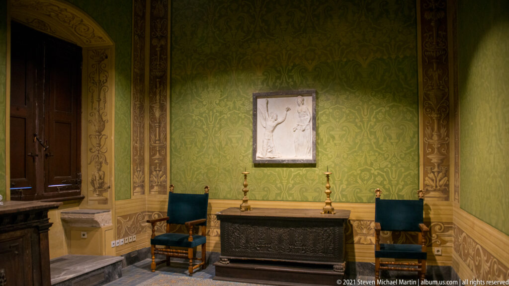 Green patterned room with dark carved chest, two high-backed chairs, candlesticks, ornamental wall borders, and a small framed relief.