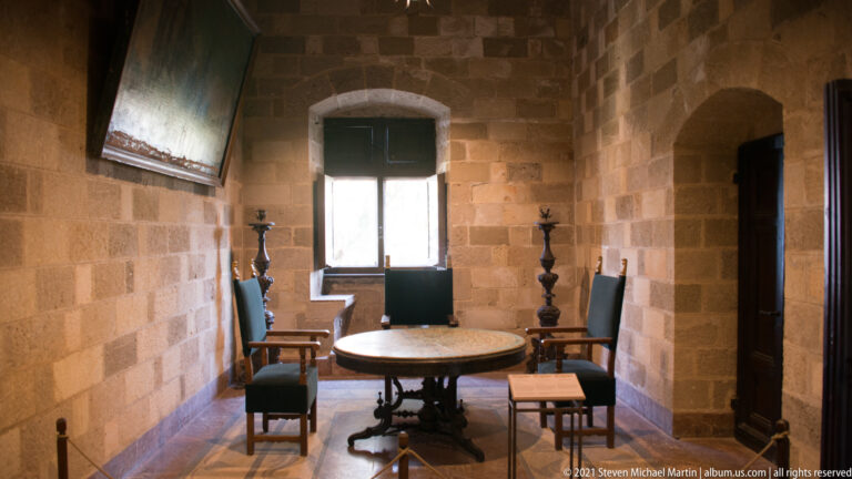 Small ashlar-stone chamber with round wooden table, high-backed chairs, tall candlesticks, bright window, and arched doorway.