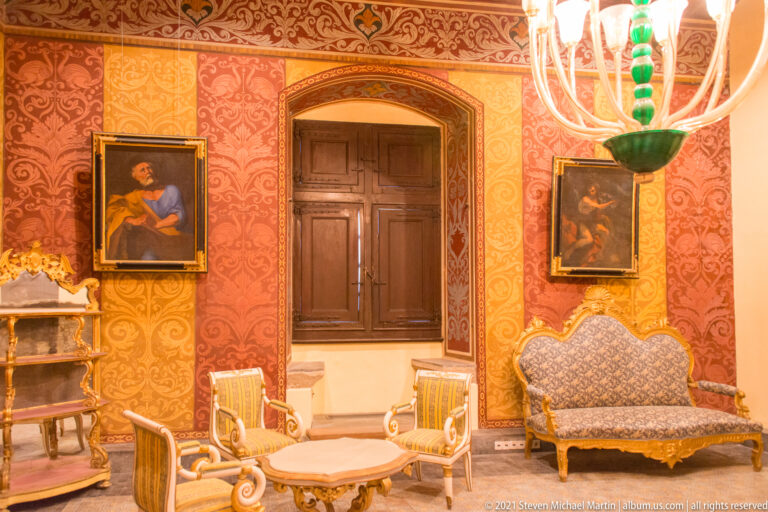 Red patterned salon with gilded chairs and sofa, two framed paintings, central shuttered window, and green glass chandelier.