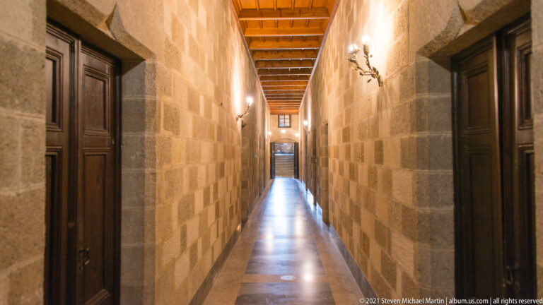 Long palace corridor with stone block walls, dark wooden doors, wall sconces, timber ceiling, and polished floor.