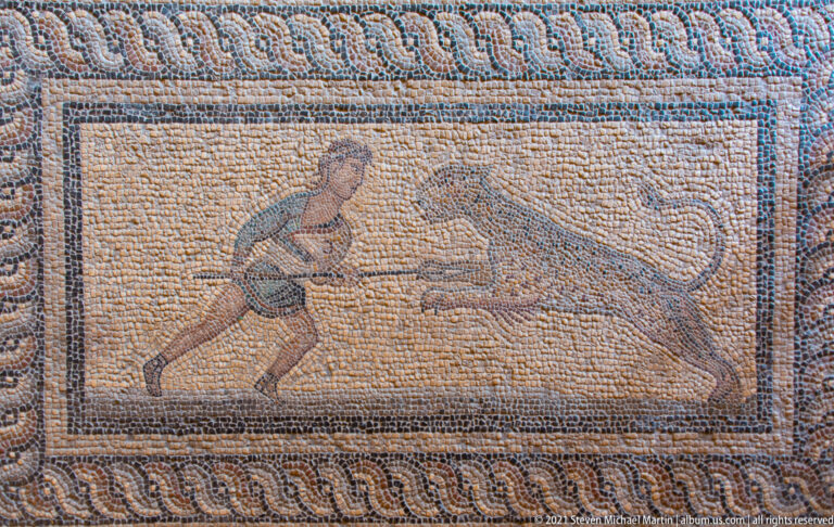 Ancient mosaic panel with a youthful figure confronting a charging bull, enclosed by a repeating wave-pattern border.