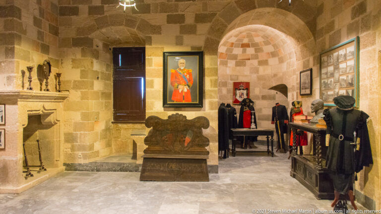 Stone museum room with mannequins in historic-style clothing, framed portraits, carved chest, arched recess, and display furnishings.