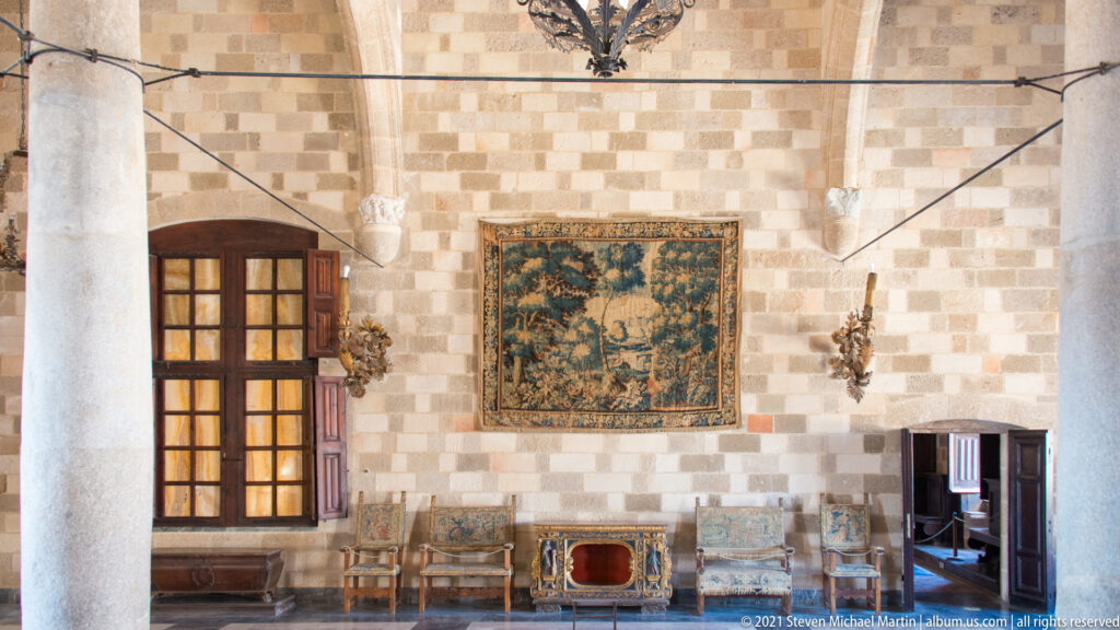 Large tapestry above upholstered chairs and small furnishings on a checker-pattern stone wall between tall wooden windows and a column.
