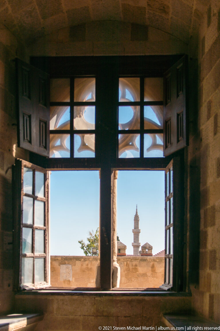 Open dark wooden casement window in a thick stone wall framing blue sky, sunlit roofline, and a slender exterior tower.