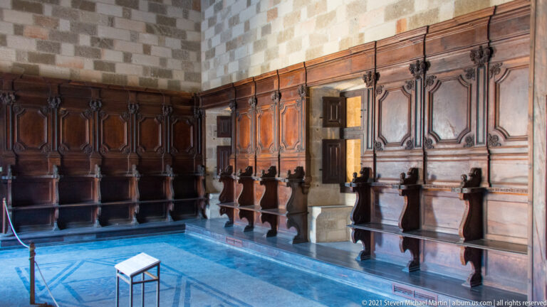 Room corner lined with dark carved wooden paneling and built-in high-backed benches above a blue patterned floor.