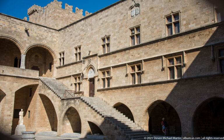 Stone staircase and arcades in the palace courtyard, with rows of mullioned windows across the sunlit upper façade.