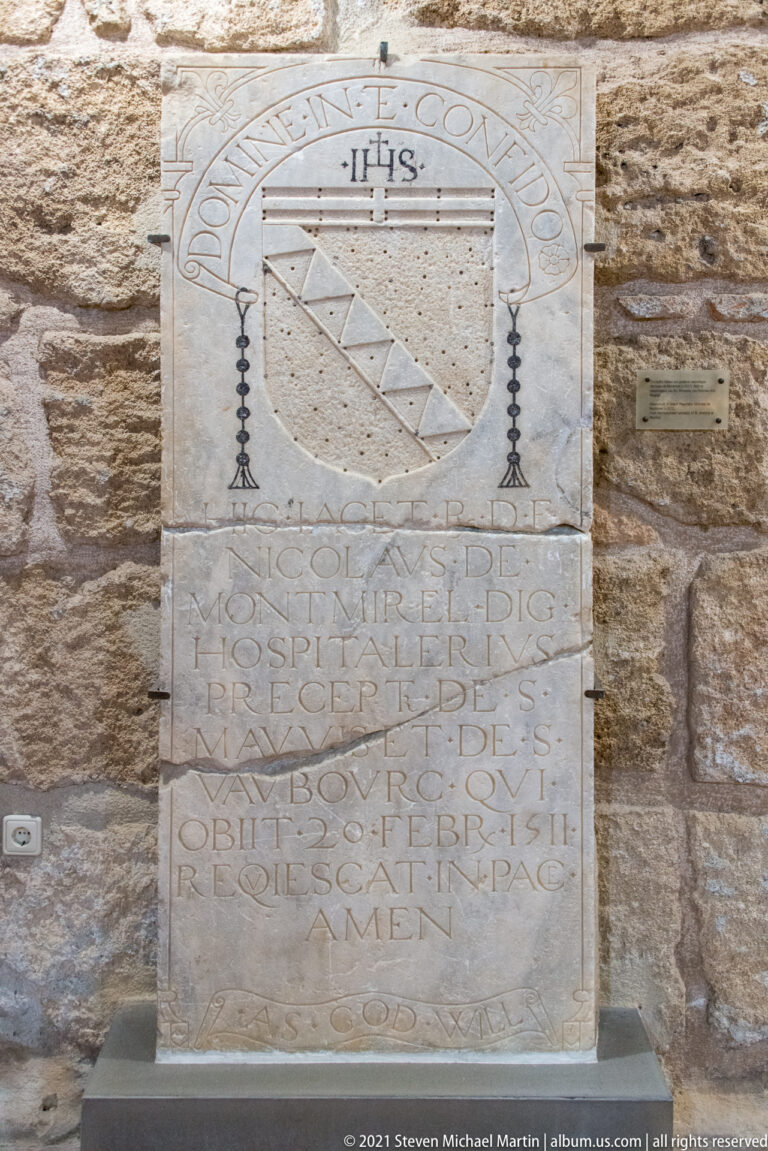 Rectangular inscribed stone plaque with a heraldic shield and Latin lettering mounted in a rough stone wall.