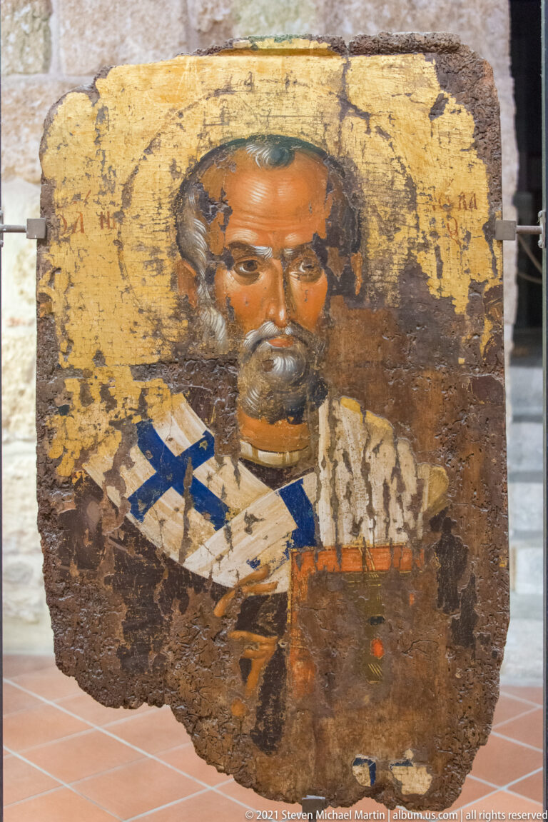 Weathered gold-ground icon of a frontal bearded bishop saint holding a decorated book, with worn Greek lettering around the head.