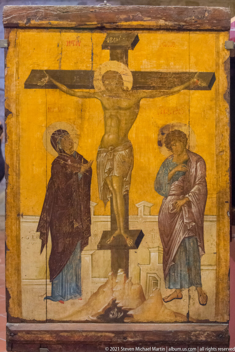 Historic Crucifixion painting with Christ on the cross between two haloed figures on a worn ochre background.