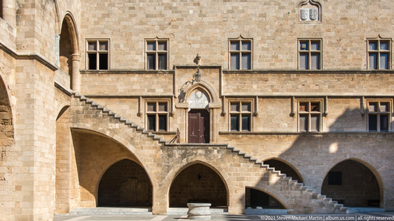 Arcaded stone courtyard façade with an exterior staircase, pointed doorway, mullioned windows, and a circular basin below.