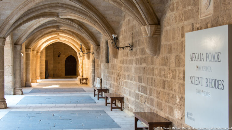 Long vaulted stone corridor with pointed arches, benches, wall sconces, and an exhibition panel reading Ancient Rhodes.